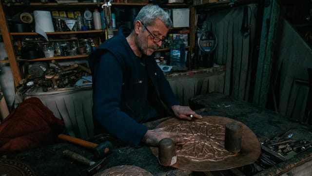 Elderly craftsman working meticulously on copper art in a traditional workshop.