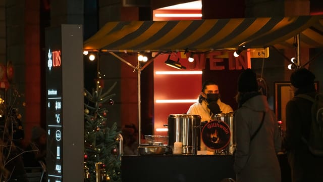 Nighttime Christmas market stall with vendor serving hot Gluhwein to visitors under festive lights.