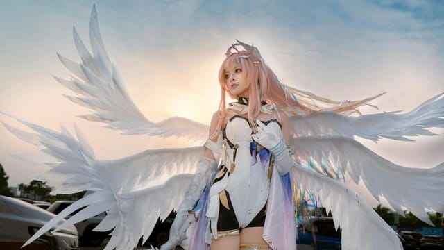 A cosplayer dressed as an angel with large wings stands outdoors at sunset.
