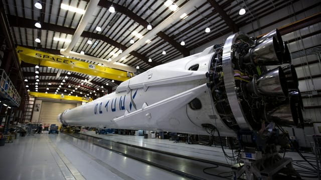 A SpaceX Falcon rocket displayed in a spacious hangar under bright industrial lights.