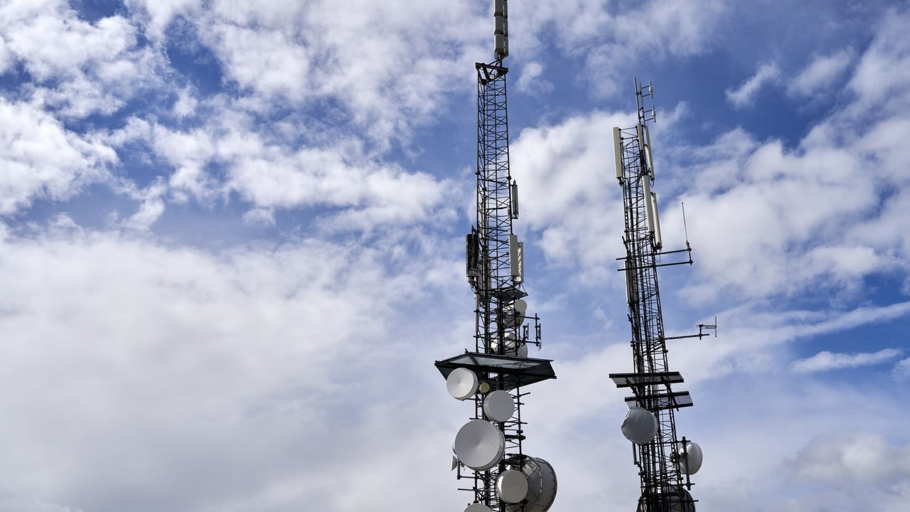 Telecommunication towers with antennas and dishes under a bright cloudy sky.