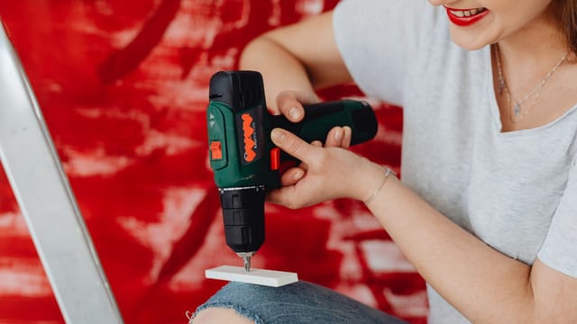 A smiling woman using a cordless drill, engaged in a home renovation project with a red background.
