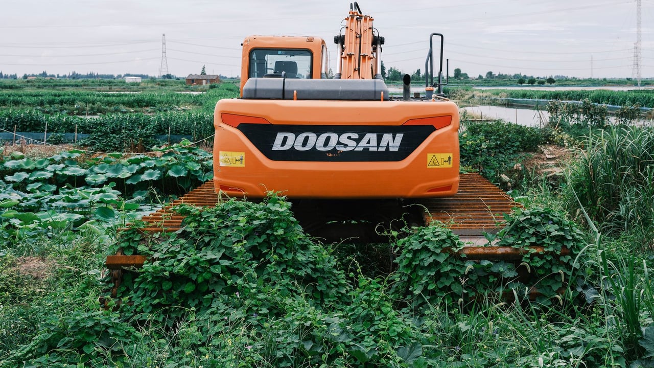 Bright orange excavator working in a lush green agricultural field under a cloudy sky.