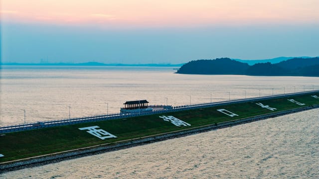A serene lakeside scene with a dam, pavilion, and hilly backdrop during sunset.