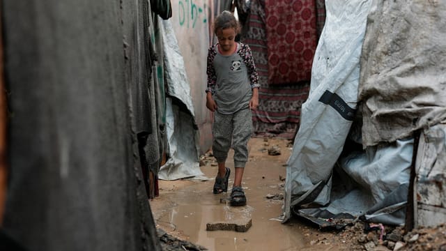 A young girl navigates a muddy path in a Gaza refugee camp, surrounded by makeshift shelters.