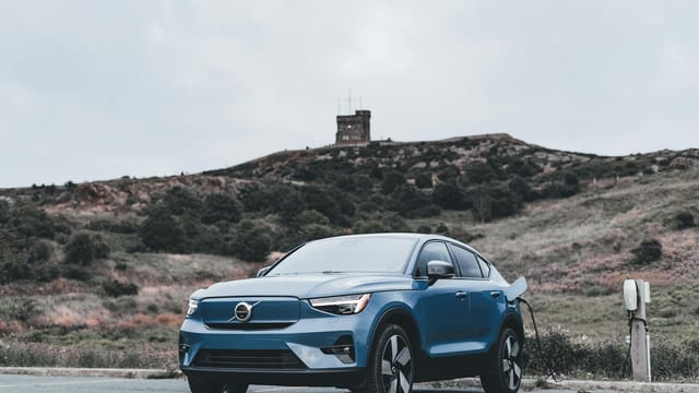 A blue Volvo electric car charging in a scenic outdoor location with a hill in the background.