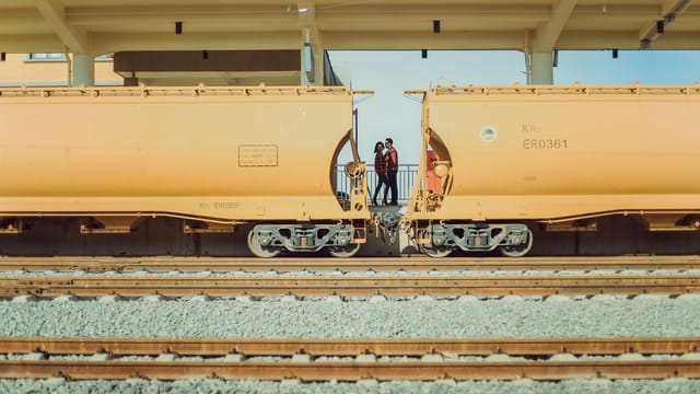 Two individuals pose between connected cargo trains at a sunlit railway station.