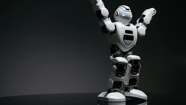 Close-up studio shot of a white robot toy with LED eyes raised in victory on a gray background.