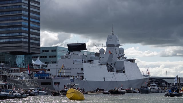 A large naval warship anchored near a modern cityscape under dramatic cloudy skies.