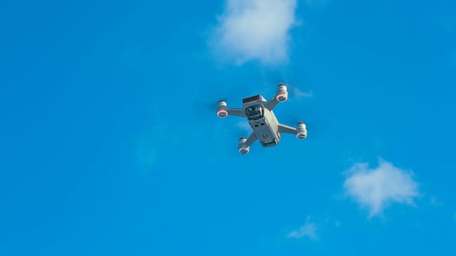 Aerial view of a drone cruising in a bright blue sky with scattered clouds.