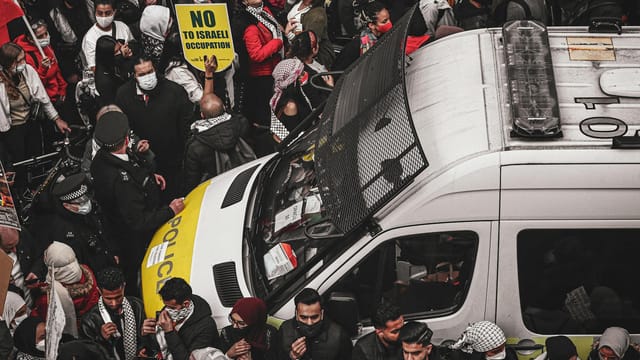 A crowd of protesters surrounding a police van holding signs during a demonstration against Israeli occupation.