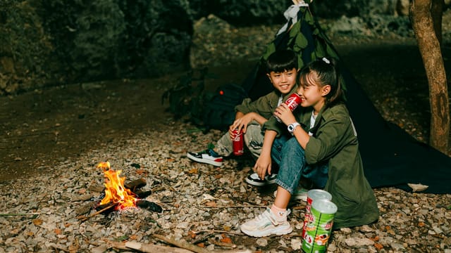 Two children sitting by a campfire, enjoying drinks and snacks in a forest setting.