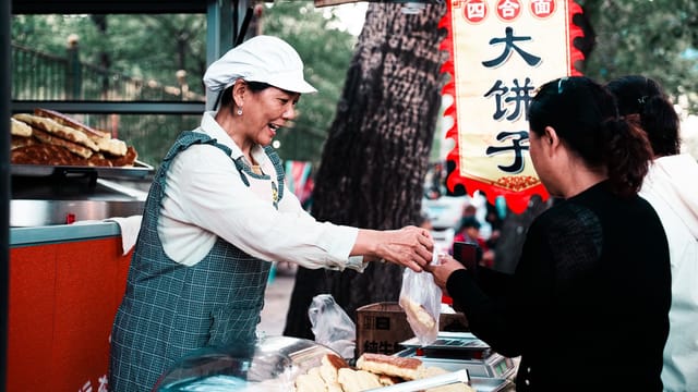 A cheerful street food vendor serving customers at an outdoor stall with traditional Chinese pastries.