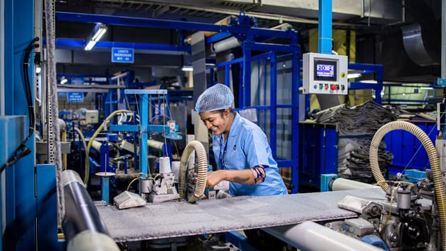Indian female factory worker smiling while operating complex textile machinery in an industrial setting.