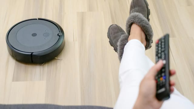 A person relaxing on a couch with a robotic vacuum cleaning the hardwood floor.