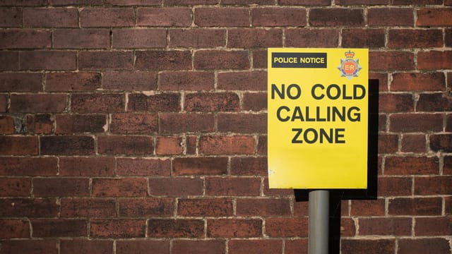 Yellow police notice sign stating 'NO COLD CALLING ZONE' against brick wall.