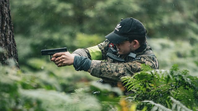 Illustrative image: A soldier in camouflage aims a pistol among ferns during rainfall, capturing a military adventure scene.