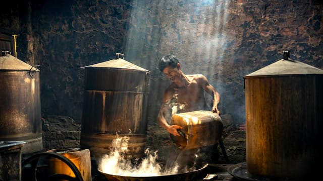 A man works in a traditional distillery, surrounded by steam and sunlight beams.