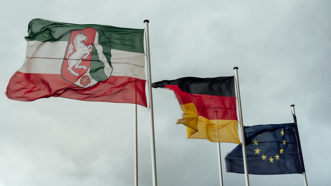 Flags of North Rhine-Westphalia, Germany, and the EU waving under a cloudy sky.