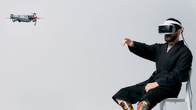 A man with prosthetic legs engages in virtual reality with a drone hovering nearby in a studio setting.
