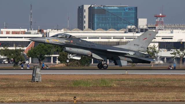 A military fighter jet in motion taking off from a runway with a cityscape background.