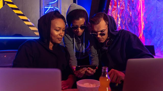 People in hoodies gathered in a cyber-themed bar using technology devices at night.