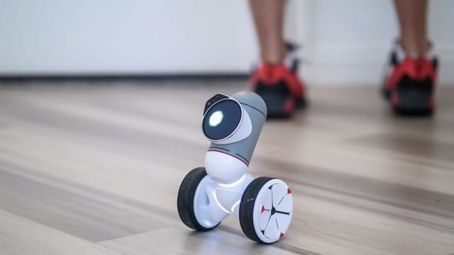 A close-up of a modern wheeled robot on a wooden floor with legs in the background.