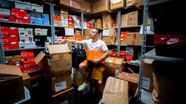 A man organizing various shoe boxes in a cluttered store room filled with shelves and cartons.