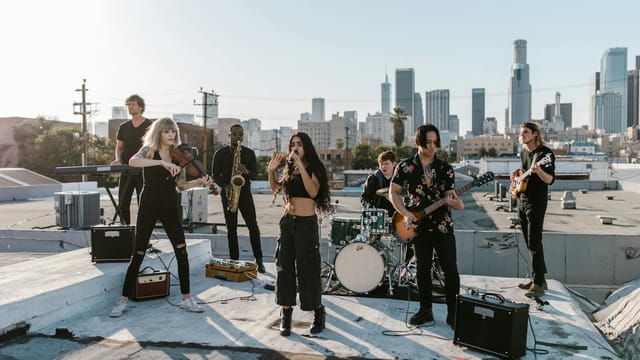 A diverse indie band performs energetically on a rooftop with a stunning Los Angeles skyline backdrop.
