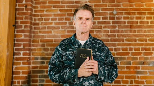 A senior U.S. Navy veteran holding a Holy Bible against a brick wall background.