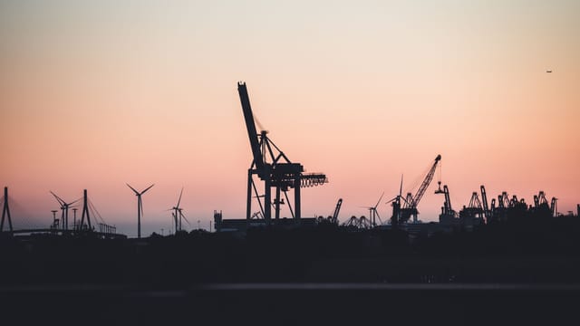Silhouetted cranes and wind turbines at the port of Hamburg during sunset, highlighting industrial beauty.