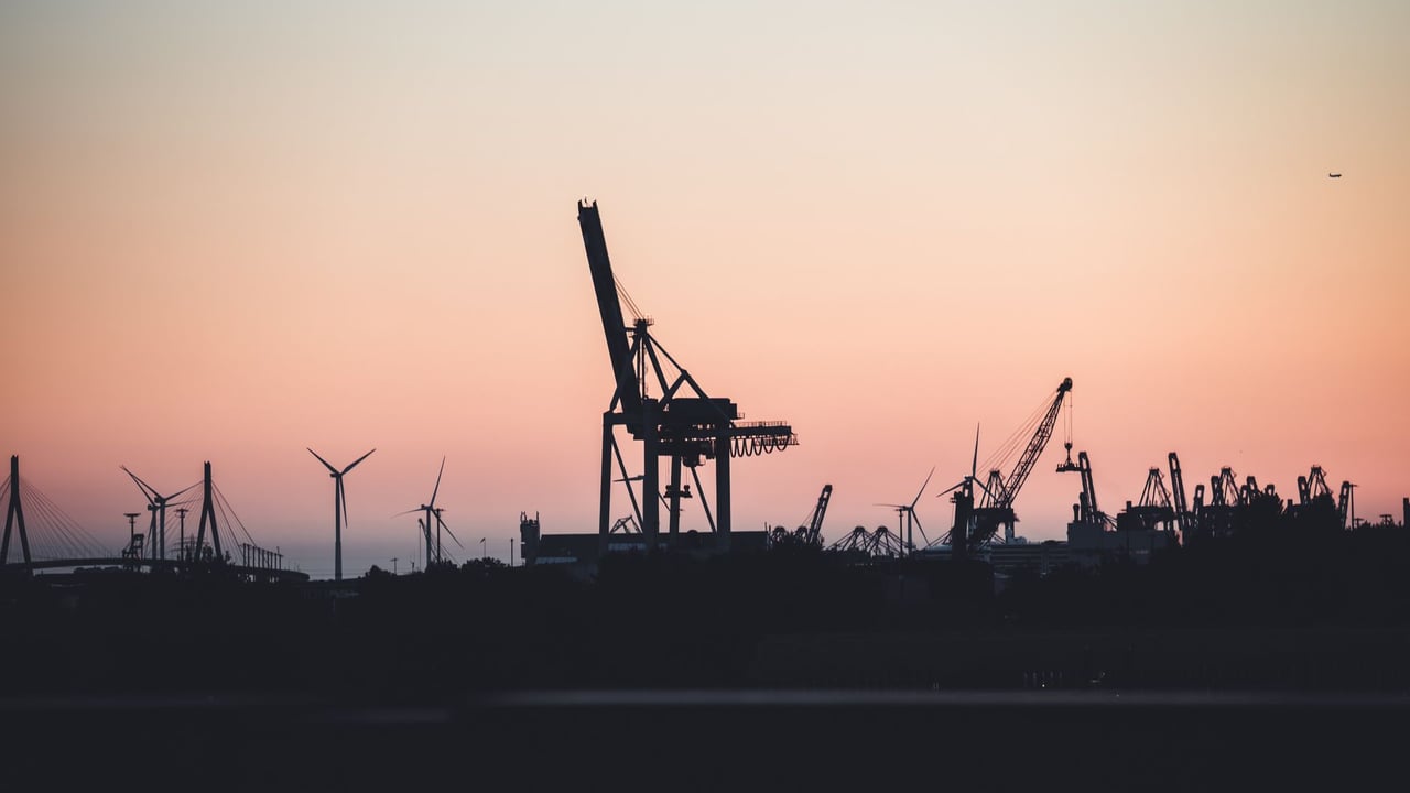 Silhouetted cranes and wind turbines at the port of Hamburg during sunset, highlighting industrial beauty.