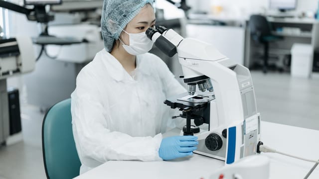 A scientist in protective gear examining samples with a microscope in a modern laboratory setting.