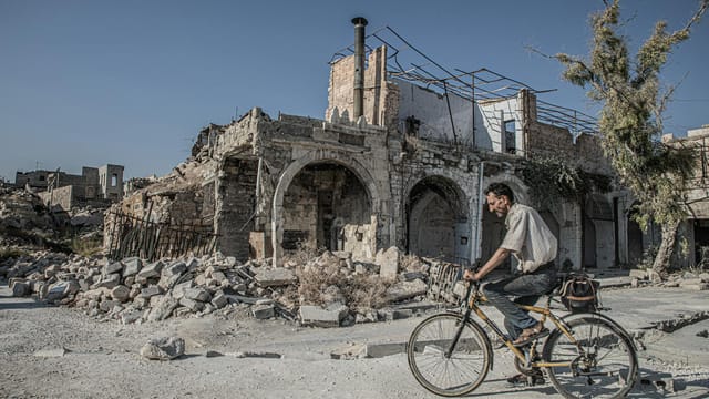 A man rides a bicycle past the ruins of Aleppo, symbolizing resilience amidst destruction.