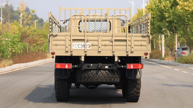 A military transport vehicle captured from behind on a quiet road, surrounded by trees.