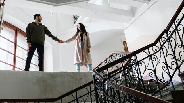 A couple romantically holds hands while standing on an ornate staircase indoors, exuding a warm and classic vibe.