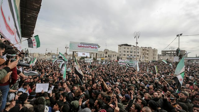 Aerial view of a large political protest in Idlib, Syria, showcasing numerous protesters holding flags and signs.