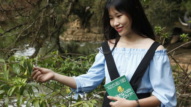Smiling woman in a park holding a book, enjoying a sunny day outdoors.
