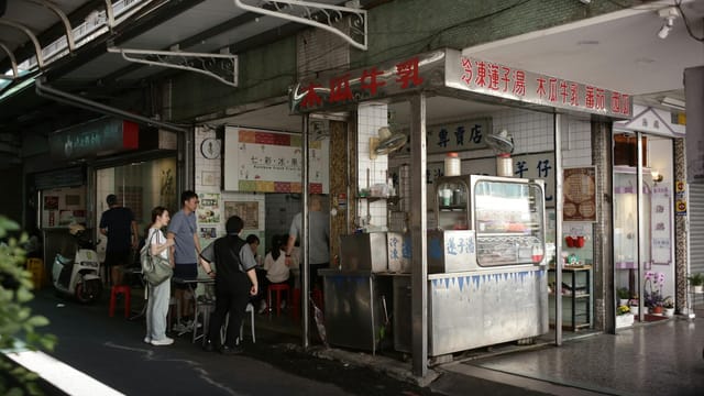 An Asian street food stall with people enjoying drinks under an awning in daylight.