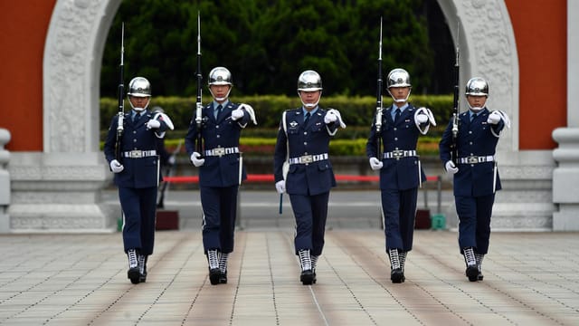 Five uniformed military personnel marching in formation during a ceremonial parade outdoors.
