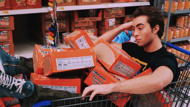 A man lounging in a shopping cart filled with Maruchan ramen in a supermarket aisle.