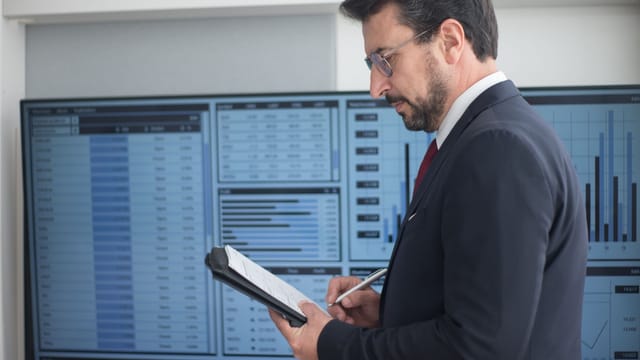Businessman in a suit analyzing data analytics on large screens, taking notes.