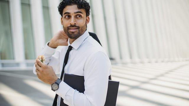 Businessman in formal attire eating a sandwich outdoors with a modern background.