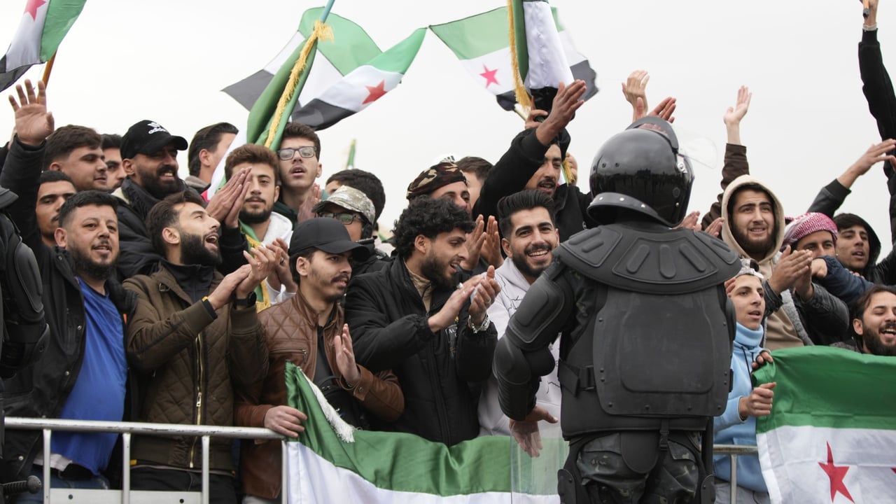 A group of men holding Syrian flags rallying around an armored officer.