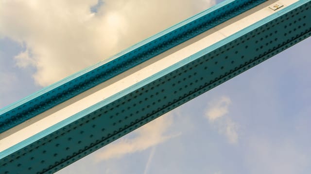 Close-up view of a blue metal bridge beam against a cloudy sky, showcasing architectural design.