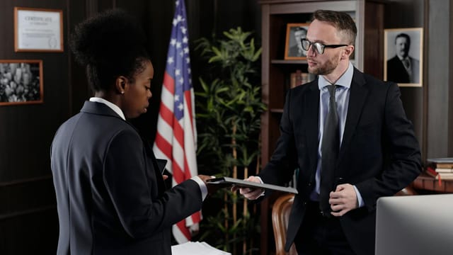 Business professionals engage in a meeting in a formal office environment with an American flag in the background.