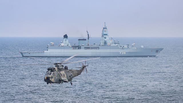 A military helicopter flies over the sea near a German warship, F219.