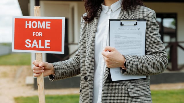 A real estate agent holding a home for sale sign and clipboard outside a property.
