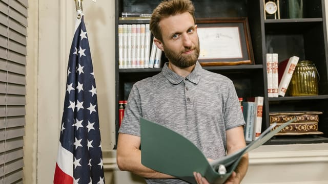 Business professional standing confidently in office holding documents near American flag.