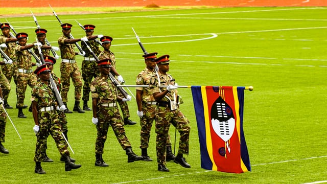 Group of Swazi soldiers in uniform participating in a ceremonial march with a national flag.